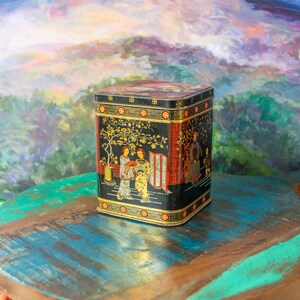 Op de afbeelding: Een rechthoekige zwarte metalen opbergdoos met gouden en rode illustraties van figuren in traditionele kleding onder een boom. De container heeft een decoratief geometrisch randpatroon langs de boven- en onderkant.