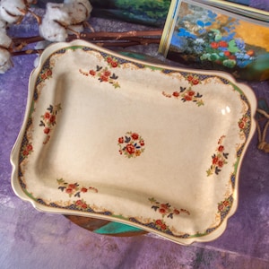 May include: A rectangular ceramic serving tray with a cream-colored base, featuring a central floral cluster and smaller floral motifs along the scalloped edges. The border is decorated with a blue and gold lattice pattern and green trim.