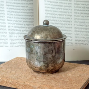 Op de afbeelding: Een kleine, cilindervormige metalen container met een afgerond deksel en een centrale knop als handvat. Het oppervlak van het vat vertoont een verweerde, doffe patina met gevlekte grijze en zilveren tinten, met tekenen van oxidatie en slijtage.