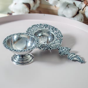 May include: A silver-toned metal tea strainer set consisting of a bowl-shaped stand with a pedestal base and a matching perforated strainer with an ornate, decorative handle featuring intricate scrollwork and floral relief patterns throughout.