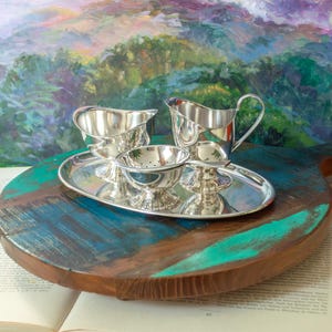 May include: A silver-toned tea service set consisting of a creamer, a sugar bowl, and a tea strainer, all arranged on a matching oval silver-toned tray. Each piece features a polished, reflective metallic finish and a pedestal base.