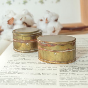 May include: Two small, round, brass containers with a weathered, tarnished patina and visible copper-toned oxidation along the center seams. Each lid is flat and smooth, and the sides feature a single small, centered metal rivet detail.