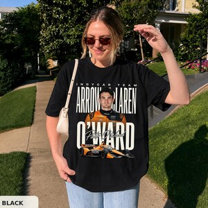 Może przedstawiać: Czarny t-shirt z krótkim rękawem z nadrukiem graficznym kierowcy IndyCar Pato O'Warda w pomarańczowym kombinezonie wyścigowym, pomarańczowym samochodem wyścigowym oraz napisem "INDYCAR TEAM ARROW MCLAREN O'WARD" białymi, pogrubionymi i kursywnymi literami.