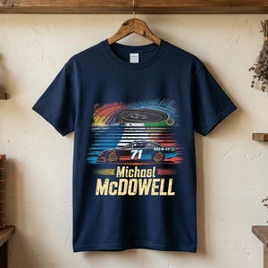 May include: A navy blue short-sleeve t-shirt featuring a graphic print of a race car with the number 71 on the side, a racetrack scene above it, and the text Michael McDOWELL printed in large yellow letters at the bottom of the design.