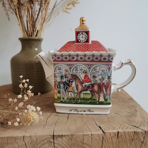 May include: A square ceramic teapot featuring a red roof with a clock tower lid, gold trim, and a colorful illustration of a horse race scene with spectators. The base of the teapot displays the text A Day at the Races in black script lettering.