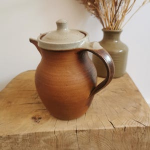 May include: A ceramic teapot with a rounded body, featuring a warm terracotta-colored base that transitions into a darker brown near the handle and spout. It includes a matching lid with a light beige glaze and a curved handle on the side.