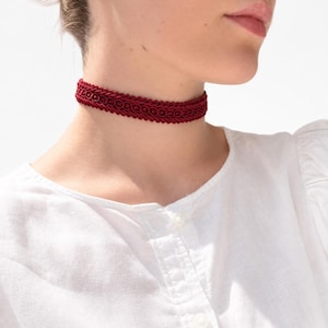 Könnte beinhalten: Eine dunkelrote Halskette im Choker-Stil mit einem gewebten, geflochtenen Besatz und einem sich wiederholenden Muster. Das Accessoire liegt eng am Hals an und zeigt eine strukturierte, kordelartige Optik mit komplexen Schlaufen.