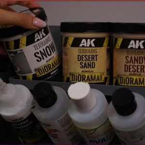 May include: Three cylindrical jars of AK Terrains acrylic diorama paste labeled Snow, Desert Sand, and Sandy Desert, featuring black lids and yellow hazard-striped labels. Below them are several clear plastic squeeze bottles with white caps.