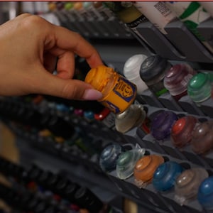 Puede incluir: Un estante de almacenamiento escalonado de plástico gris que contiene numerosos botes de pintura pequeños con tapas de colores. Se está retirando un bote de pintura naranja del estante, que cuenta con múltiples ranuras inclinadas.