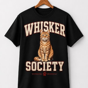 Peut inclure: Un t-shirt noir à manches courtes avec un motif représentant un chat tigré orange assis. Au-dessus du chat, le texte "WHISKER" et en dessous le texte "SOCIETY" sont inscrits en rouge vieilli, avec une petite empreinte de patte rouge en bas.