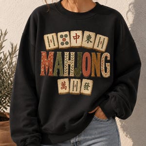 Peut inclure: Un sweat-shirt noir à col rond arborant une impression graphique de tuiles de Mahjong disposées en rangées au-dessus et au-dessous du mot MAHJONG en grandes lettres décoratives multicolores. Les tuiles présentent divers symboles et caractères chinois.
