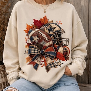 Peut inclure: Un sweat-shirt à col rond couleur crème arborant un motif central composé d'un ballon de football, d'un casque et d'une citrouille décorés de motifs à carreaux et d'étoiles. Le design inclut des feuilles d'automne, des baies et un grand nœud.