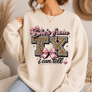 May include: A cream-coloured crewneck sweatshirt featuring a graphic print with the text "She's from TX i can tell". The design includes large block letters with a leopard print pattern, accented by pink bows, small stars, and a heart icon.