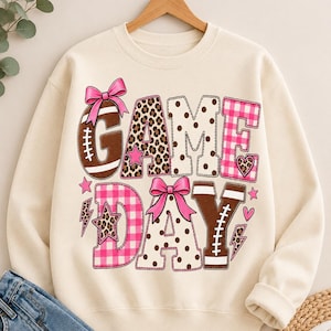 Peut inclure: Un sweat-shirt à col rond couleur crème avec l'inscription GAME DAY en grandes lettres à motifs variés. Le design inclut des lettres marron style ballon de football, du vichy rose, des motifs léopard et pois, avec des nœuds, étoiles et cœurs roses.