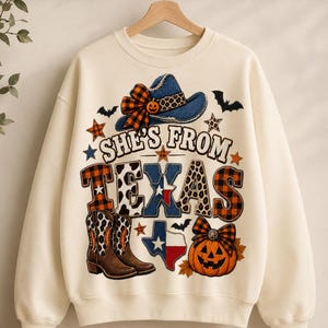 Peut inclure: Un sweat-shirt à col rond couleur crème avec une impression graphique représentant un chapeau de cow-boy bleu, une citrouille, des bottes de cow-boy et le texte "SHE'S FROM TEXAS". Le motif inclut des carreaux, des taches de léopard, des chauves-souris, des étoiles et la forme du drapeau du Texas.