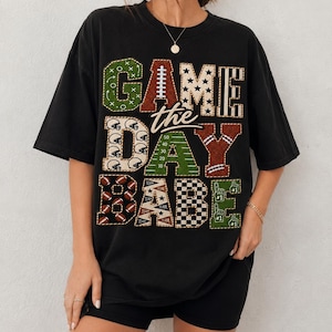Peut inclure: Un t-shirt noir à manches courtes avec un grand imprimé graphique portant le texte GAME DAY BABE. Les lettres sont remplies de divers motifs sur le thème du football, notamment des ballons, des casques, des étoiles et des marquages de terrain.