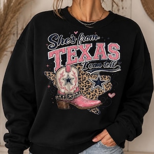 Peut inclure: Un sweat-shirt noir à col rond avec une impression graphique portant le texte "She's from TEXAS I can tell". Le motif comprend une botte de cow-boy rose et blanche, une carte du Texas à imprimé léopard, une étoile bleue et des cœurs roses.