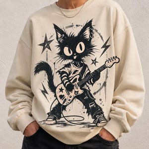 May include: A cream-colored crewneck sweatshirt featuring a black graphic illustration of a cat playing an electric guitar. The cat has large eyes, a tongue sticking out, and wears a t-shirt, ripped pants, and sneakers, surrounded by stars and lightning bolts.