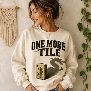 May include: A cream-coloured crewneck sweatshirt featuring the black text ONE MORE TILE above a graphic of a mahjong bamboo tile and a dark dragon silhouette. The long-sleeved garment has a ribbed collar, cuffs, and hem, providing a relaxed fit.