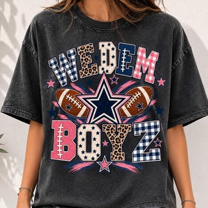 May include: A charcoal gray short-sleeve t-shirt featuring the text WEDEM BOYZ in colorful patterned block letters. The design includes a central navy blue star, two brown footballs, and small pink stars on a washed-out black fabric background.