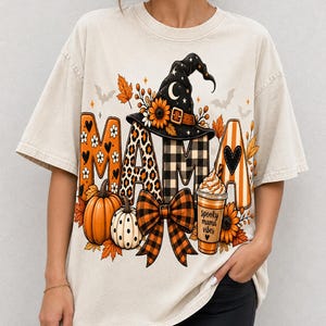 Peut inclure: Un t-shirt beige à manches courtes avec une impression graphique du mot MAMA dans divers motifs, un chapeau de sorcière noir avec des tournesols, des citrouilles, un nœud à carreaux et une tasse de café avec le texte spooky mama vibes.