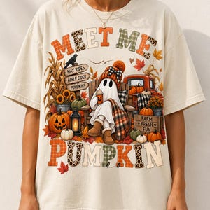 Peut inclure: Un t-shirt couleur crème avec une impression graphique portant le texte "MEET ME PUMPKIN". Le motif représente un fantôme portant un bonnet et des bottes, assis sur une chaise, entouré de citrouilles, d'un camion rouge et de tournesols.