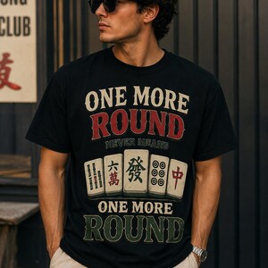 Peut inclure: Un t-shirt noir à manches courtes avec le texte ONE MORE ROUND NEVER MEANS ONE MORE ROUND en lettres crème et rouges, centré au-dessus et au-dessous d'une illustration graphique de cinq tuiles de mah-jong avec divers symboles.