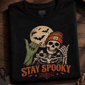 Peut inclure: Un t-shirt noir avec un motif représentant un squelette portant un bonnet orange avec un smiley, faisant un signe shaka à côté d'une main de zombie verte. Le design inclut une pleine lune avec des chauves-souris, un damier et le texte STAY SPOOKY.