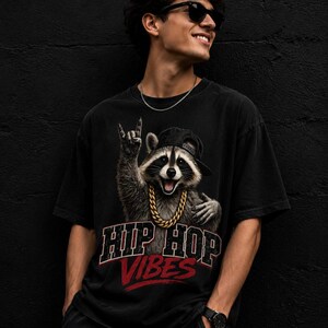 Peut inclure: Un t-shirt noir à manches courtes avec un motif représentant un raton laveur portant une casquette noire et une chaîne dorée. Le raton laveur fait un geste de la main. Sous le motif, le texte HIP HOP VIBES est écrit en lettres rouges et blanches.