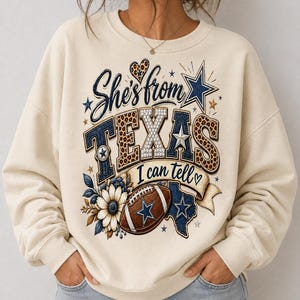 Peut inclure: Un sweat-shirt à col rond couleur crème avec une impression graphique portant le texte "She's from TEXAS I can tell". Le motif comprend un ballon de football, une carte du Texas, une fleur, des étoiles et des accents léopard en bleu, marron et blanc.