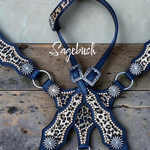 May include: A dark blue leather horse breast collar featuring leopard print inlays, silver-toned starburst conchos, and small gold-toned studs. The piece includes two silver-toned buckles and the word Sagebush written in white script across the center.