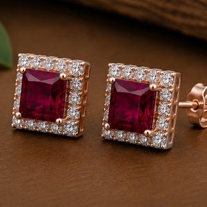 Peut inclure: Une paire de boucles d'oreilles carr&eacute;es avec une pierre centrale rouge fonc&eacute; taille princesse entour&eacute;e d'un halo de petites pierres rondes transparentes serties dans une monture en m&eacute;tal or rose avec une tige de fermeture.