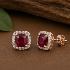 Könnte beinhalten: Ein Paar Ohrstecker aus Roségold, jeweils mit einem tiefroten Rubin im Kissenschliff in der Mitte, umgeben von einem Kranz aus kleinen, klaren, rund geschliffenen weißen Diamanten. Die Ohrringe verfügen über Standard-Steckverschlüsse.
