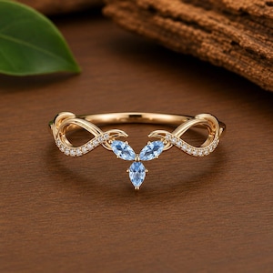 Puede incluir: Un anillo de oro con una banda en forma de infinito adornada con pequeñas piedras claras. El centro del anillo presenta tres piedras preciosas de color azul claro con corte marquesa dispuestas en racimo, creando un diseño delicado y simétrico.