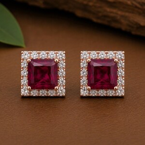 Peut inclure: Une paire de boucles d'oreilles carr&eacute;es avec une pierre centrale rouge fonc&eacute; taille princesse, entour&eacute;e d'un halo de petites pierres blanches rondes et transparentes, le tout mont&eacute; sur une structure en m&eacute;tal dor&eacute; rose.