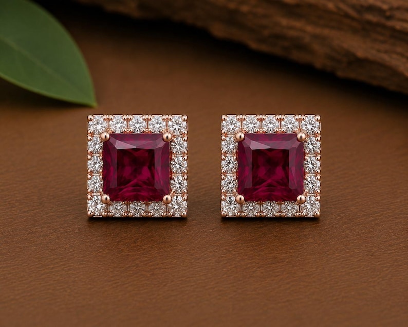 Peut inclure: Une paire de boucles d'oreilles carr&eacute;es avec une pierre centrale rouge fonc&eacute; taille princesse, entour&eacute;e d'un halo de petites pierres blanches rondes et transparentes, le tout mont&eacute; sur une structure en m&eacute;tal dor&eacute; rose.