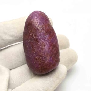 May include: A raw, tumbled ruby gemstone with a deep magenta and dark reddish-brown colour palette. The stone features an irregular, elongated oval shape with a textured, matte surface showing natural mineral inclusions and subtle crystalline facets.