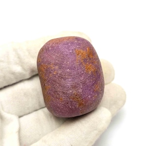 Pode incluir: Uma pedra preciosa polida e arredondada com um tom magenta e roxo profundo, apresentando manchas proeminentes de inclusões minerais terrosas de cor castanho-alaranjada na sua superfície texturizada, com uma forma natural e orgânica.