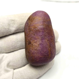 Peut inclure: Une pierre précieuse polie, lisse et arrondie, d'une teinte magenta et violette profonde, présentant des inclusions et des veines minérales brun doré irrégulières sur sa surface opaque et texturée, de forme naturelle et organique.