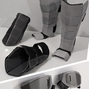 May include: A set of protective gear including two grey shin guards with rivet details, two black forearm guards with adjustable straps, and two grey wrist guards with rectangular cutouts. The pieces feature layered panels and black interior padding.