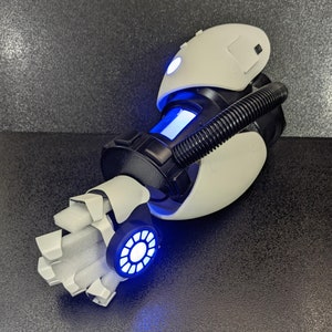 Puede incluir: Un brazo robótico blanco y negro con una luz azul. El brazo tiene una cabeza blanca redonda con forma de casco con una manguera negra unida a ella. El brazo tiene una mano blanca con forma de garra con una luz azul en el centro.