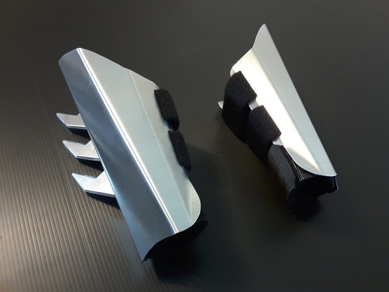 May include: A pair of silver-colored metal forearm guards with black straps. The guards have a sharp, pointed design on the front.