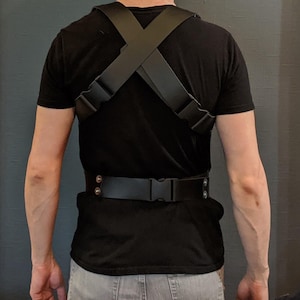 May include: A black leather harness with adjustable straps and a buckle closure. The harness is worn over a black t-shirt.