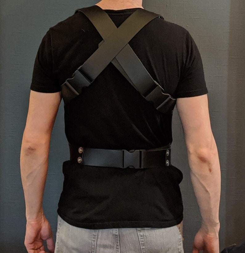 May include: A black leather harness with adjustable straps and a buckle closure. The harness is worn over a black t-shirt.