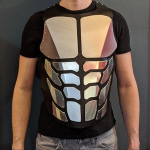 May include: A silver-coloured, segmented chest plate with a black fabric backing. The chest plate is worn over a black t-shirt.