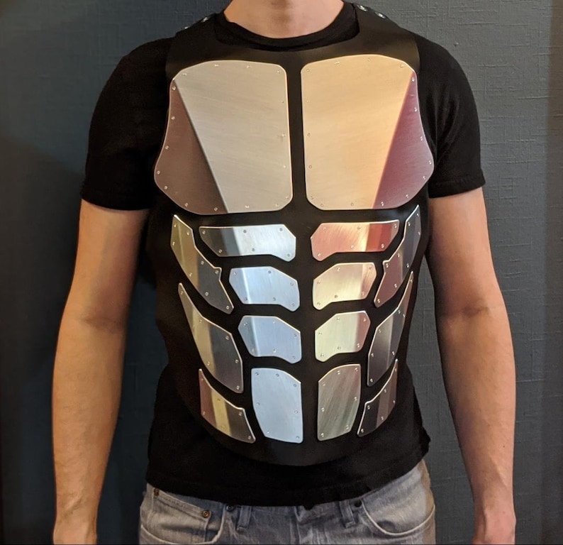 May include: A silver-coloured, segmented chest plate with a black fabric backing. The chest plate is worn over a black t-shirt.