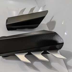 May include: A pair of black arm guards with silver claws. The guards are made of a textured material and have a sleek, modern design.