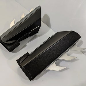 May include: A pair of black arm guards with silver blades. The guards are made of a textured material and have a strap to secure them to the arm.