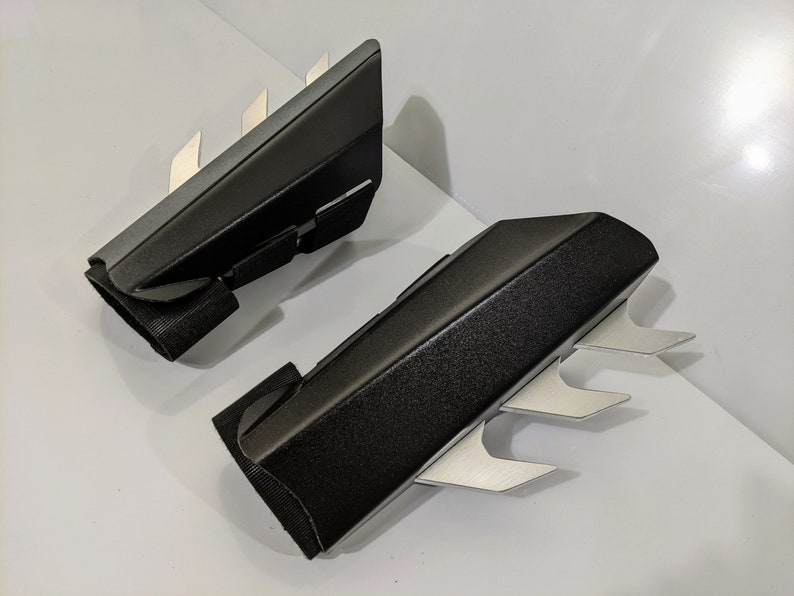 May include: A pair of black arm guards with silver blades. The guards are made of a textured material and have a strap to secure them to the arm.