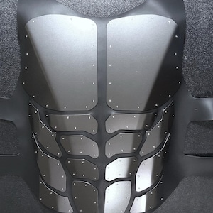 May include: A black and silver chest plate with a textured design. The chest plate is made of multiple pieces of metal that are connected by rivets.